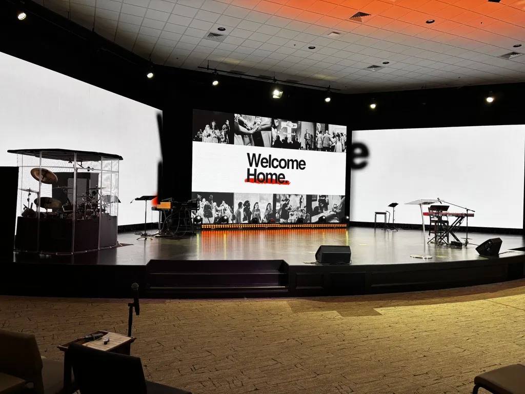 The City Sanctuary stage today: curved black stage with center stairs, a center LED wall on a lit plinth, two large angled side LED walls, drum shield stage left and keys stage right.